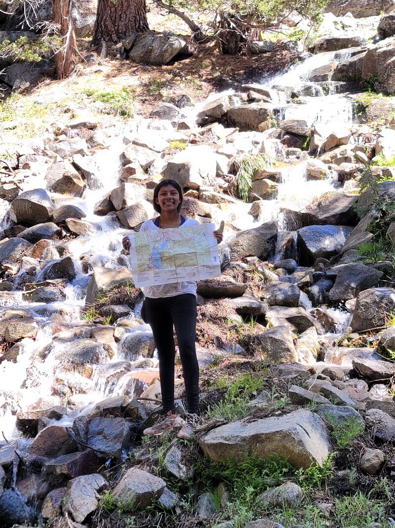 A woman stands on rocks in front of a small waterfall, holding a map. She is wearing a white shirt and dark pants. The background consists of rocks, trees, and the flowing water of the waterfall. The scene appears to be outdoors, possibly in a mountainous or natural area. The woman is smiling and seems to be enjoying her time in nature.